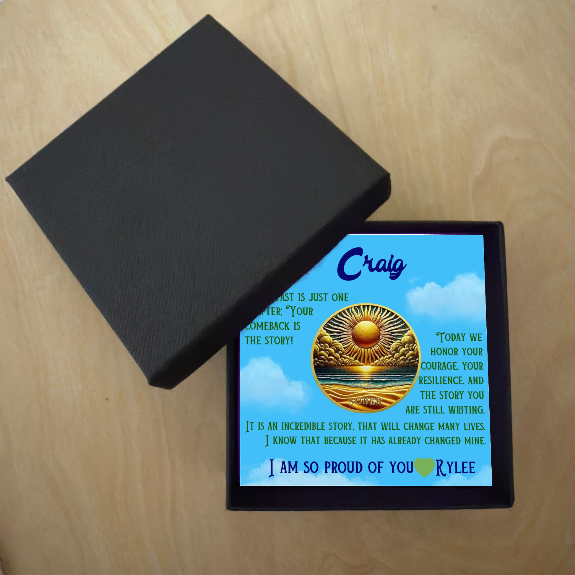 Black gift box with lid and blue greeting card featuring Craig and gold sun-over-ocean coin