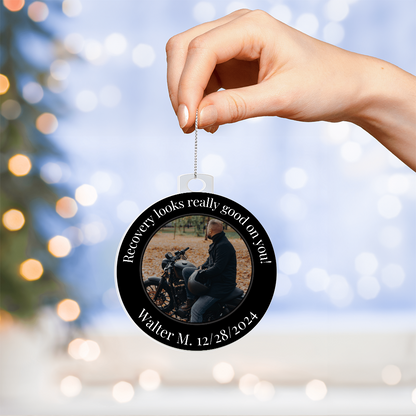 Round acrylic recovery photo ornament hanging from a silver chain in front of soft lights