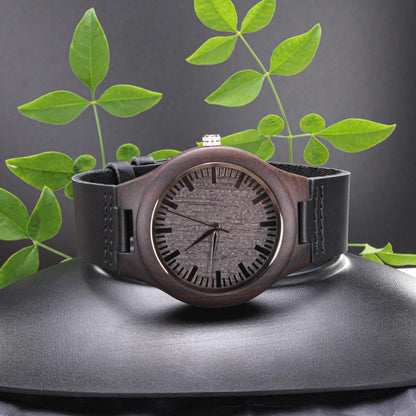 Wood watch with dark round case and black leather band, engraved round dial and three hands.