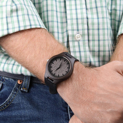 Wood watch on wrist with dark round case, black leather strap, textured dial.