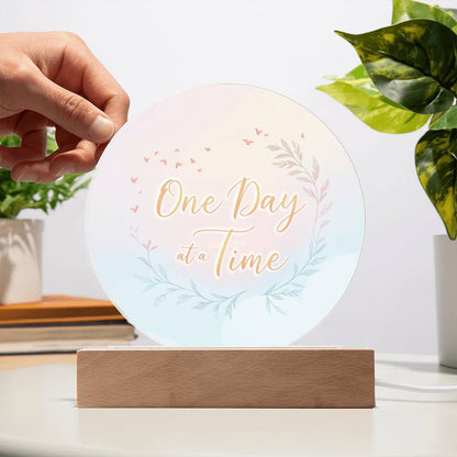 Hand holding a round acrylic LED plaque reading "One Day at a Time" on wood base