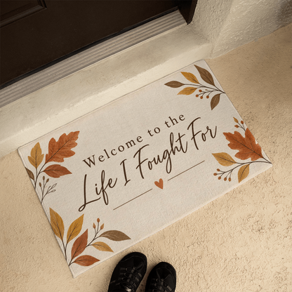 Beige doormat angled on tile, featuring leaf corners and the quote "Welcome to the Life I Fought For"