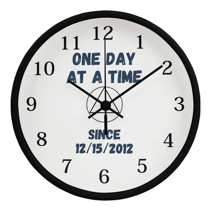 Black-framed wall clock with black numerals and blue “ONE DAY AT A TIME” text