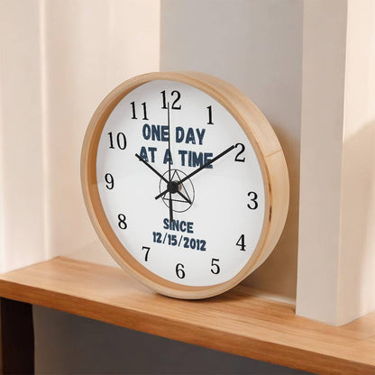 Natural wood wall clock on shelf with black hands and blue “ONE DAY AT A TIME” text