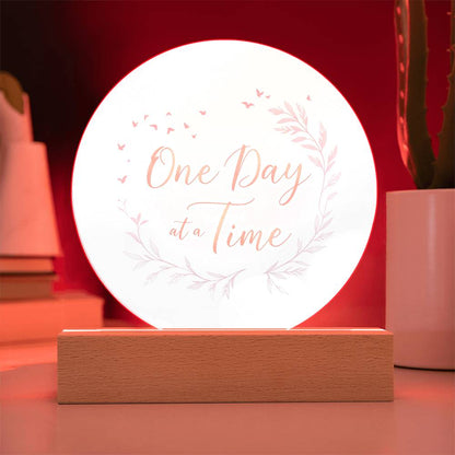 Illuminated round acrylic LED plaque sign saying "One Day at a Time" on wood base