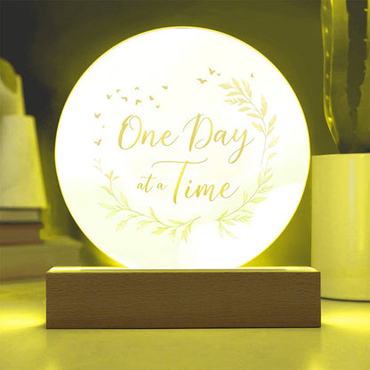 Warmly lit round acrylic LED plaque with "One Day at a Time" floral design, wood base