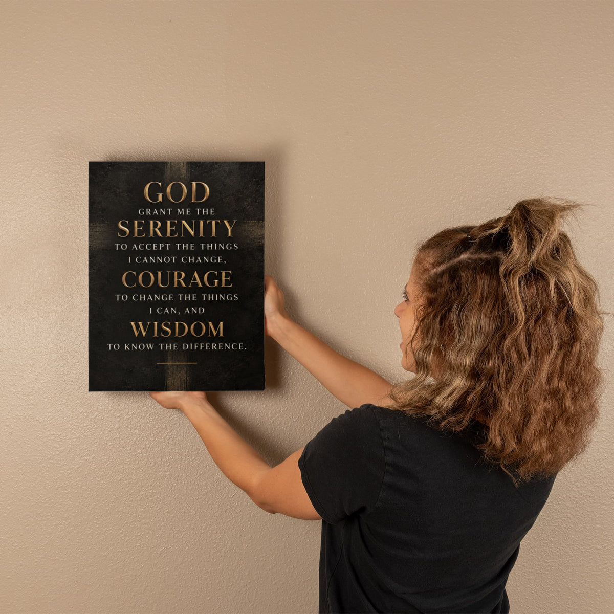Black vertical canvas wall art with gold Serenity Prayer text, held against wall.
