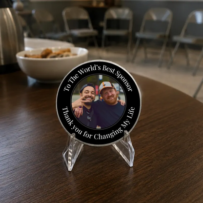Round black photo coin with silver rim on a clear stand; features two smiling men.