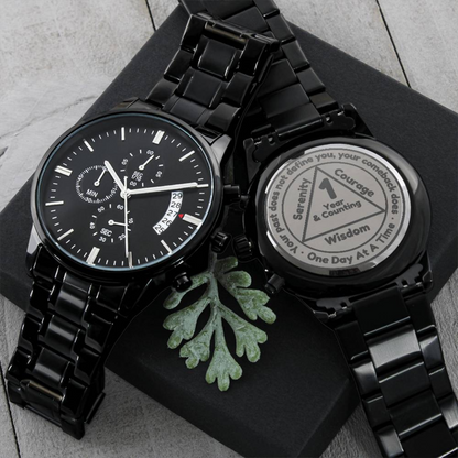 Black recovery watch with linked metal bracelet and round dial beside engraved token